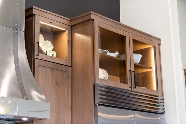 Warm wooden upper cabinets with glass-front doors and interior lighting showcase decorative ceramic pieces and tableware. Positioned beside a stainless steel range hood and refrigerator, the design blends functionality with rustic elegance.